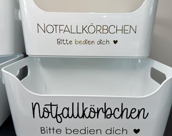 May include: White rectangular storage bins with curved handles. The bins have the text "NOTFALLKÖRBCHEN Bitte bedien dich" in gold and black lettering, with a heart symbol. The bins are stacked on top of each other.