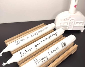 May include: White candles with black text reading "Wein & Kerzenschein", "Let's go camping", and "Happy Camper". The candles are on a wooden holder, with a white campervan-shaped candle holder in the background.