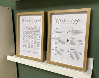 May include: Two framed prints with light wood frames. One print displays laundry symbols, the other offers stain removal tips. The prints are mounted on a white shelf against a green wall. The text on the prints is in German.