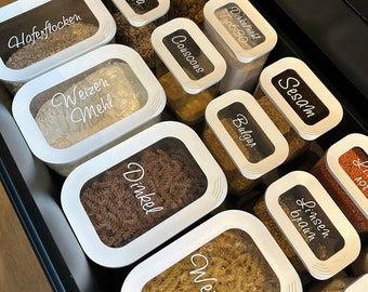 May include: A white drawer containing clear containers filled with different grains and pasta. The containers are labelled with handwritten labels in German, including "Haferflocken", "Walnüsse", "Dinkel Mehl", "Typ 630", "Sesam", "Quinoa", "Couscous", "Bulgur", "Linsen braun", "Weizen Mehl", "Dinkel", and "Weizen".