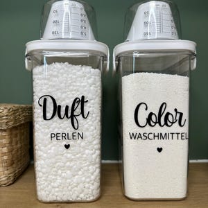 Könnte beinhalten: Zwei transparente, rechteckige Behälter mit weißen Deckeln und Messbechern. Ein Behälter ist mit "Duft PERLEN" beschriftet und mit weißen Perlen gefüllt. Der andere ist mit "Color WASCHMITTEL" beschriftet und mit weißem Pulver gefüllt. Wäschekammer-Organisation.