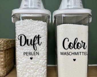 May include: Two clear, rectangular containers with white lids and measuring cups. One container is labelled "Duft PERLEN" and filled with white beads. The other is labelled "Color WASCHMITTEL" and filled with white powder. Laundry room storage.