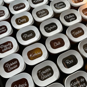 May include: A close-up of a white spice rack with various spices labeled in black lettering. The labels include "Oregano", "Muskat", "Kümmel", "Pfeffer schwarz", "Curry", "Pizza", "Paprika edelsuß", "Kurkuma", "Tomaten Gewürz", "Gulasch", "Steak Gewürz", "Wacholder beepen", "Lorbeer Blätter", "Piri Piri", "Thymian", "Nelken ganz", "Tandori", "Chili", "Reis", "Kräuter der Provence", "Zimt", "Bohnenkraut", "Leb".