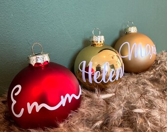 May include: Three glass Christmas baubles with names written on them. The baubles are red, gold, and gold. The names are "Emma", "Helen", and "Mary".