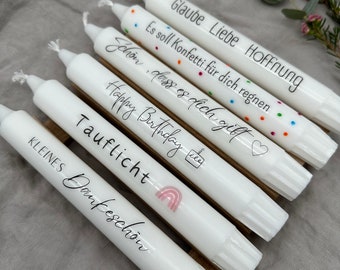 May include: Six white candles with black script and colourful accents. The candles have phrases like "Happy Birthday" and "Glaube Liebe Hoffnung". A rainbow and cake icon are also visible. The candles are arranged on a wooden surface.