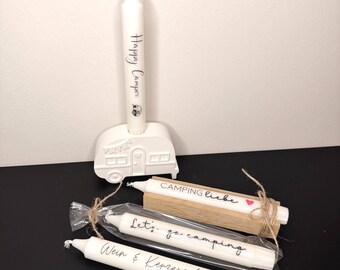 May include: White candles with black script, including "Happy Camper" and "Let's go camping." One candle is in a white camper-shaped holder. Another candle is on a wooden block with "Camping Liebe" and a heart. The candles are tied with twine.