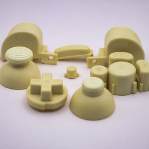 May include: A set of beige replacement buttons for a video game controller. The buttons include a directional pad, four face buttons labeled A, B, R, and L, and two analog stick caps.