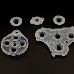 May include: Five clear silicone buttons for a video game controller. The buttons are various shapes and sizes, with some having multiple holes. One button has the number "38" printed on it.
