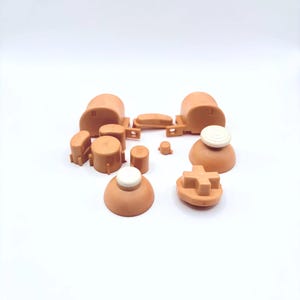 Pode incluir: Um conjunto de bot&otilde;es de substitui&ccedil;&atilde;o para um controlador de videogame. Os bot&otilde;es s&atilde;o feitos de pl&aacute;stico marrom claro e incluem um direcional, quatro bot&otilde;es frontais, dois bot&otilde;es de ombro e duas tampas de joystick anal&oacute;gico. Os bot&otilde;es s&atilde;o todos de formas e tamanhos diferentes.
