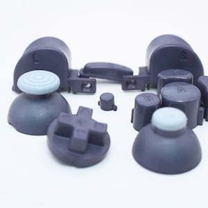 May include: A set of purple and light blue replacement buttons for a video game controller. The set includes two analog sticks, a D-pad, four face buttons, two shoulder buttons, and two triggers.