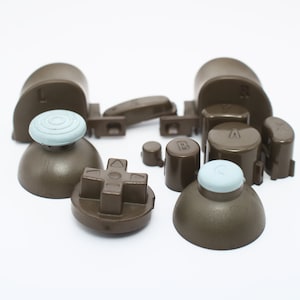 May include: A set of brown replacement buttons for a video game controller. The buttons include a directional pad, four face buttons, two analog stick caps, and two shoulder buttons. The buttons have a light blue color on the top.