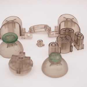May include: A set of translucent gray replacement parts for a video game controller. The set includes two face buttons, two shoulder buttons, two thumbsticks, and a d-pad.