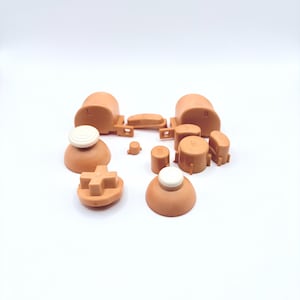 May include: A set of replacement buttons for a video game controller. The buttons are made of a light brown plastic and have a variety of shapes and sizes. The set includes a directional pad, four face buttons, two shoulder buttons, and two analog stick caps.