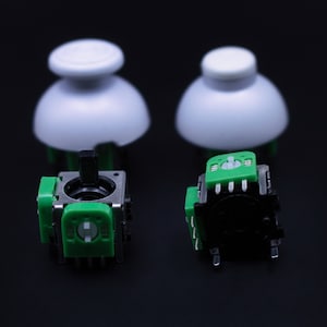 May include: Two black and green potentiometers with a white knob on each side. The potentiometers are on a black surface.