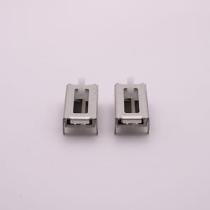 May include: Two silver-toned slide switches with white plastic actuators. The switches are rectangular and have a small opening in the centre.