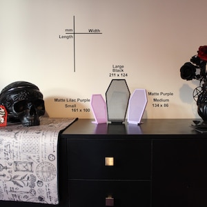 Op de afbeelding: Een zwarte schedelsculptuur, tashanger en doodskistvormige decoratiestukken worden tentoongesteld op een zwarte kast. De tashanger meet 89 x 54 mm. De doodskistdecoratie omvat een groot zwart stuk (211 x 124 mm) en twee matte paarse stukken.