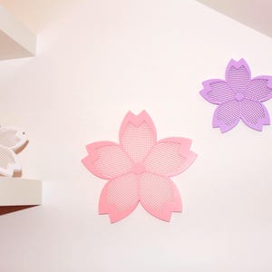 Puede incluir: Tres decoraciones de pared con forma de flor en tonos rosa y morado. Una es blanca con un peque&ntilde;o elemento decorativo. Las flores tienen un patr&oacute;n de malla y est&aacute;n montadas en una pared blanca, creando un tema floral decorativo.