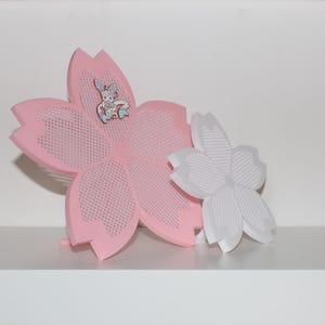 Puede incluir: Una decoraci&oacute;n rosa en forma de flor con un peque&ntilde;o alfiler de conejo adjunto. Una flor blanca m&aacute;s peque&ntilde;a est&aacute; al lado de la rosa. La flor rosa tiene un dise&ntilde;o perforado. Los art&iacute;culos est&aacute;n en un estante blanco.