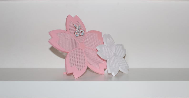 Puede incluir: Una decoraci&oacute;n rosa en forma de flor con un peque&ntilde;o alfiler de conejo adjunto. Una flor blanca m&aacute;s peque&ntilde;a est&aacute; al lado de la rosa. La flor rosa tiene un dise&ntilde;o perforado. Los art&iacute;culos est&aacute;n en un estante blanco.