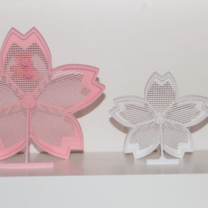 Puede incluir: Dos objetos decorativos con forma de flor. Uno es rosa, el otro blanco. Ambos tienen un dise&ntilde;o calado delicado y est&aacute;n montados sobre peque&ntilde;os soportes. La flor rosa es m&aacute;s grande que la blanca. Los objetos se exhiben en una estanter&iacute;a blanca.