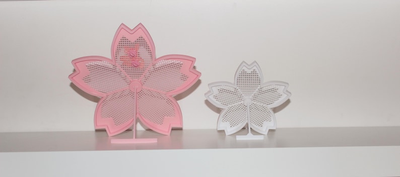 Puede incluir: Dos objetos decorativos con forma de flor. Uno es rosa, el otro blanco. Ambos tienen un dise&ntilde;o calado delicado y est&aacute;n montados sobre peque&ntilde;os soportes. La flor rosa es m&aacute;s grande que la blanca. Los objetos se exhiben en una estanter&iacute;a blanca.