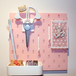 Pode incluir: Um organizador de painel perfurado rosa com vários artigos de papelaria. Inclui tesouras brancas com um detalhe de urso, um lápis azul claro, uma bandeja branca com fita washi e um cartão decorativo com uma ilustração de coelho.