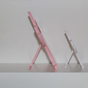 Puede incluir: Dos peque&ntilde;os marcos de fotos de madera. Uno es rosa claro, el otro blanco. Ambos tienen un dise&ntilde;o sencillo tipo caballete, con un marco rectangular y una pata de soporte en la parte trasera. Se exhiben en una estanter&iacute;a blanca.
