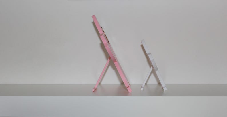 Puede incluir: Dos peque&ntilde;os marcos de fotos de madera. Uno es rosa claro, el otro blanco. Ambos tienen un dise&ntilde;o sencillo tipo caballete, con un marco rectangular y una pata de soporte en la parte trasera. Se exhiben en una estanter&iacute;a blanca.
