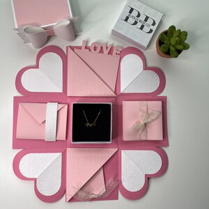 May include: Pink explosion box with a white heart design. The box has a necklace inside, a pink envelope, and a pink gift box with a white ribbon. The box is labeled "LOVE" and "BUOUX BOX CO."