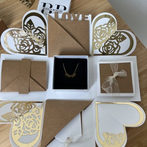 May include: A white and gold explosion box with a heart-shaped design. The box is open to reveal a small black box with a gold necklace inside. The necklace has a nameplate pendant that says "EMMA". The box also has a small brown card with a white ribbon on it.