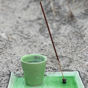 May include: A green ceramic cup with a brown incense stick burning on a green rectangular tray. The tray has a black border.