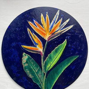 May include: A round painting of a bird of paradise flower. The flower has orange and yellow petals with blue accents, set against a dark blue background. Green leaves and gold accents complete the floral artwork.