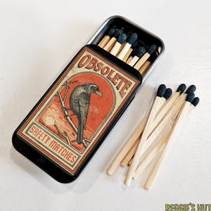 May include: A black metal matchbox with the word "OBSOLETE" and "SAFETY MATCHES" printed on the front. The box features a vintage design with a crow illustration. Several matches with blue tips are visible.