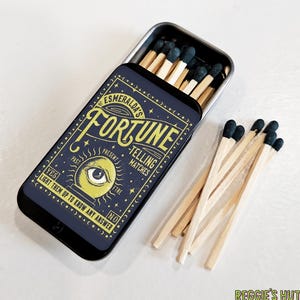 May include: A navy blue metal matchbox with the text "ESMERALDA'S FORTUNE - TELLING MATCHES" and a yellow eye graphic. The box is open, revealing wooden matches with dark green tips. A small pile of matches sits beside the box.