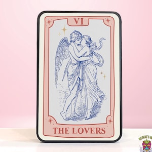 May include: A tarot card featuring a blue and white illustration of two figures embracing, with angel wings. The card is framed in a pink border with the words "THE LOVERS" and the Roman numeral "VI". The card has a black border.