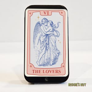 May include: A black rectangular object with a tarot card design. The card features a blue illustration of two figures embracing, with the words "THE LOVERS" in pink. The card has a pink border and the number "VI" at the top.
