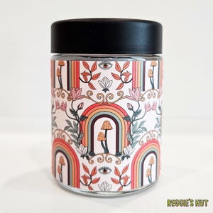 May include: A glass jar with a black lid, featuring a colorful design of rainbows, mushrooms, and floral elements. The jar's design includes a repeating pattern of rainbows, mushrooms, and stylized eyes. The text "REGGIE'S HUT" is visible at the bottom.
