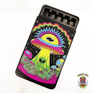 May include: Black metal case with a colorful psychedelic design featuring a large mushroom, an alien, and other smaller mushrooms. The case has a black interior with multiple compartments. The text "Reggie's Hut" is visible.