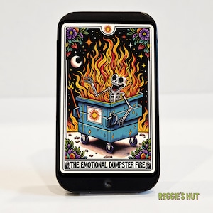May include: Black phone case featuring a tarot card design. The card depicts a skeleton engulfed in flames inside a blue dumpster, with the text "THE EMOTIONAL DUMPSTER FIRE". The design includes a crescent moon, stars, and floral accents.