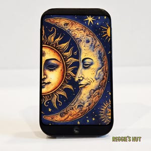 May include: Black rectangular phone case featuring a celestial design. The design includes a sun and a crescent moon with a face, set against a dark blue starry background. The sun is orange and yellow, and the moon is yellow.