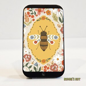 May include: A black phone case featuring a bee illustration within a yellow oval, surrounded by a floral pattern. The bee has brown and yellow stripes, with white and blue flower accents on its wings. The case has the text "Reggie's Hut".