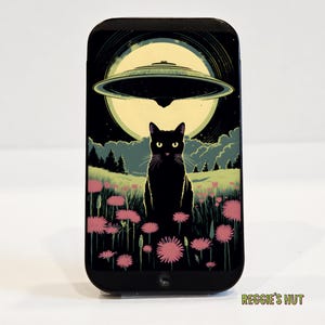 May include: Black rectangular phone case featuring a black cat in a field of pink flowers under a full moon and a UFO. The case has the text "Reggie's Hut" at the bottom.