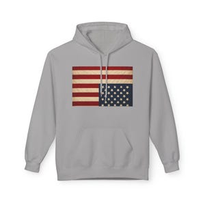 May include: A light grey hoodie with a distressed American flag design. The flag graphic features red and white stripes and a blue square with white stars. The hoodie has a front pocket and a hood with a drawstring.