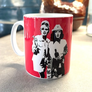 Puede incluir: Una taza de cerámica roja con asa blanca presenta una ilustración en blanco y negro de dos personas sosteniendo bebidas. Las personas visten ropa elegante y tienen un aspecto retro.