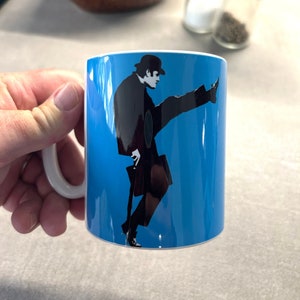 Puede incluir: Una taza de cerámica blanca con fondo azul y una silueta negra de una persona con sombrero y traje que levanta la pierna. La persona sostiene un bastón.