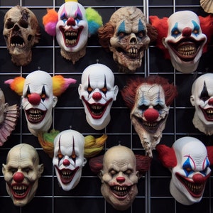 Puede incluir: Una colección de 11 máscaras de payaso espeluznantes con diversas expresiones y colores, incluyendo rojo, azul, verde y amarillo. Las máscaras están colgadas en una rejilla de metal negro.