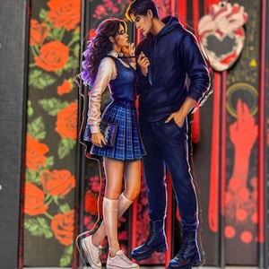 Puede incluir: Una figura de pie de acrílico transparente con una pareja en una pose romántica. La mujer lleva una falda a cuadros azul, una camisa blanca y zapatillas deportivas. El hombre viste una sudadera con capucha y pantalones azul marino. El fondo incluye diseños florales y en forma de corazón.