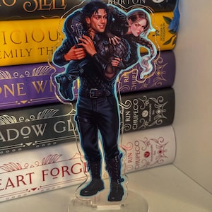 May include: A clear acrylic stand-up featuring a detailed illustration of two characters. The male character is carrying the female character on his back. The artwork is set against a backdrop of colorful books.