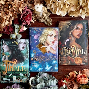 May include: Three hardback books with illustrated covers, titled "Finale", "Legendary", and "Caraval". The covers feature portraits of women, with floral and decorative elements. The books are arranged on a wooden surface, surrounded by flowers.