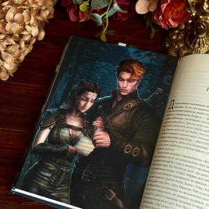 Puede incluir: Un libro abierto con un marcador de página, con una ilustración de fantasía de un hombre y una mujer con ropa oscura. La portada representa una escena de bosque. Rosas rojas y otras flores están en el fondo.